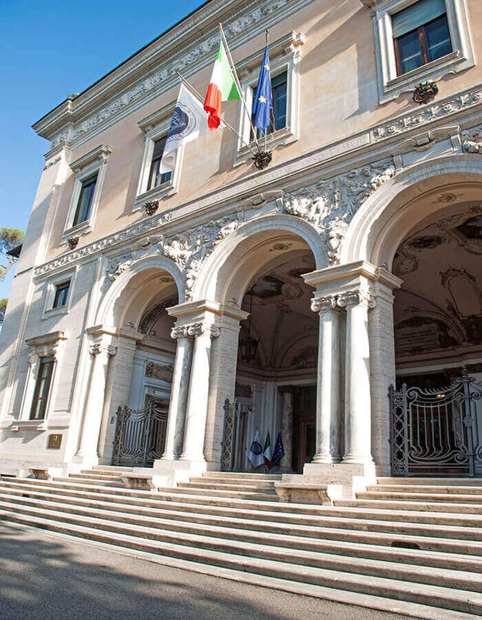 Vista della monumentale facciata e scalinata di Villa Lubin a Roma, sede del CNEL, con archi, colonne e bandiere.
