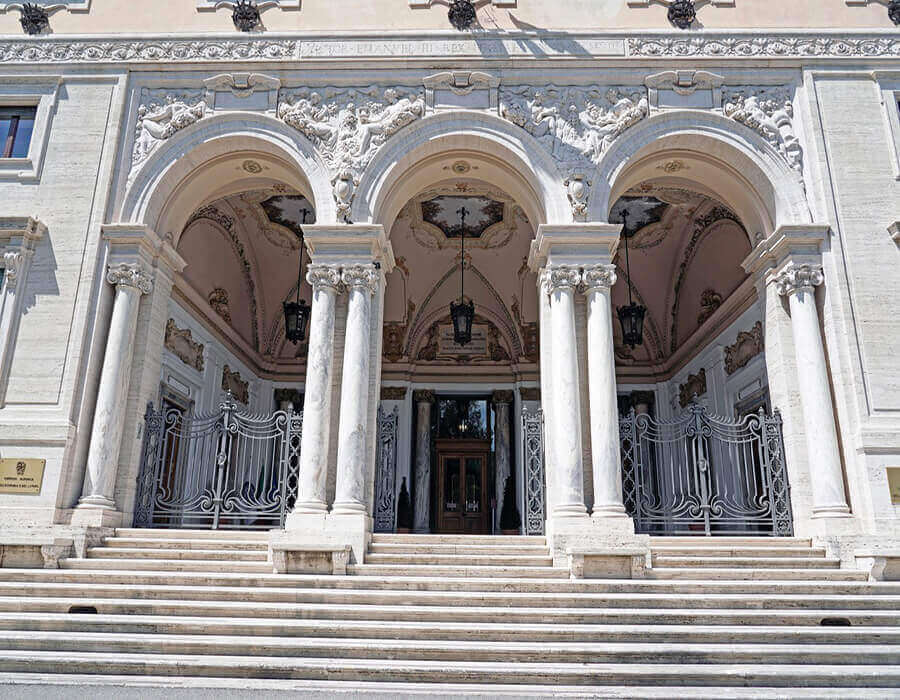Veduta frontale dell'ingresso di Villa Lubin a Roma. Una grande scalinata in marmo conduce a un portico con tre archi e colonne classiche. Sono visibili decorazioni in bassorilievo, cancelli in ferro battuto, lanterne e la porta d'ingresso in legno.
