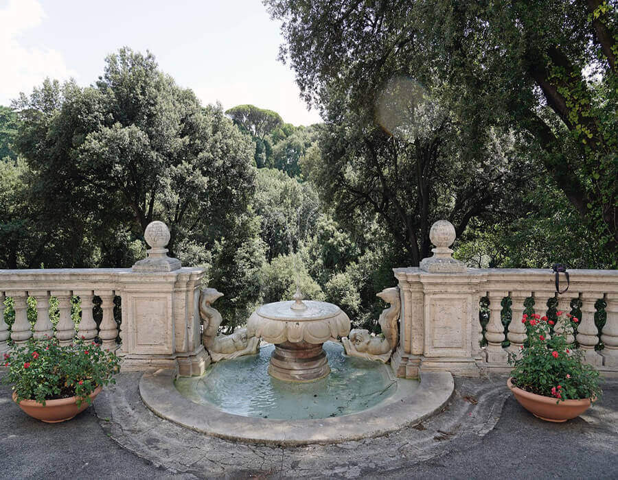 Particolare della terrazza di Villa Lubin a Roma. Una fontana decorativa in pietra antica con figure scolpite è al centro. È affiancata da una balaustra in pietra e da due vasi di terracotta con fiori rossi. Oltre la balaustra, una vista su un bosco