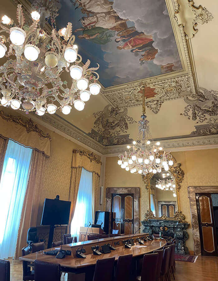 Interno di una sala riunioni a Villa Lubin a Roma, sede del CNEL. Si vedono un grande tavolo ovale in legno, lampadari in vetro di Murano, un soffitto affrescato, pareti decorate, un camino e una specchiera.