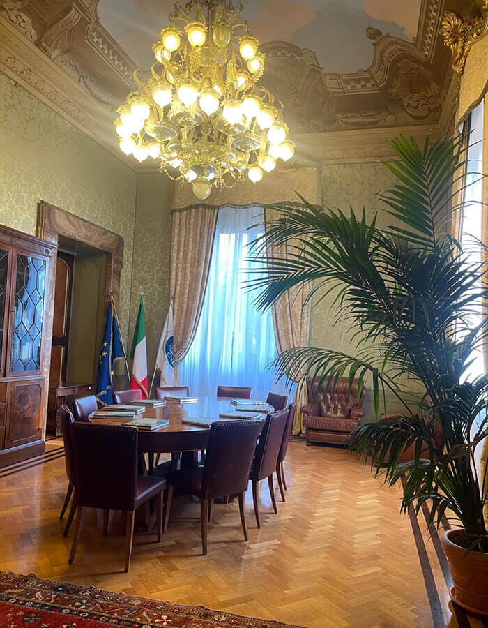Interno di una sala riunioni a Villa Lubin a Roma, sede del CNEL. Include un tavolo rotondo, sedie in pelle, un lampadario ornamentale e un soffitto affrescato. Sono presenti l'armadio, le bandiere italiana ed europea e una grande pianta.