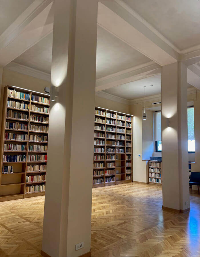 Interno della biblioteca di Villa Lubin a Roma, sede del CNEL. La sala presenta alte librerie in legno colme di libri, pavimenti in parquet a spina di pesce e pilastri strutturali bianchi.